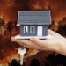 A model house rests on a hand against a backdrop of a large wildfire.