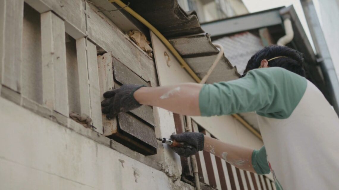 A worker uses a drill to repair wooden siding on the exterior of a house.