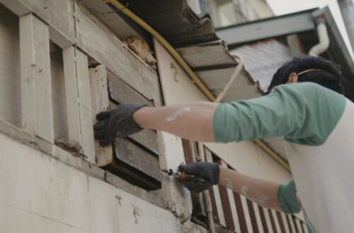 A worker uses a drill to repair wooden siding on the exterior of a house.