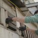 A worker uses a drill to repair wooden siding on the exterior of a house.