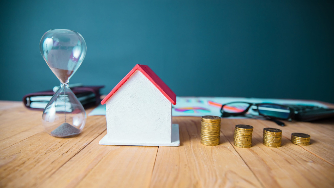 A model house sits beside an hourglass and stacks of gold coins on a desk.