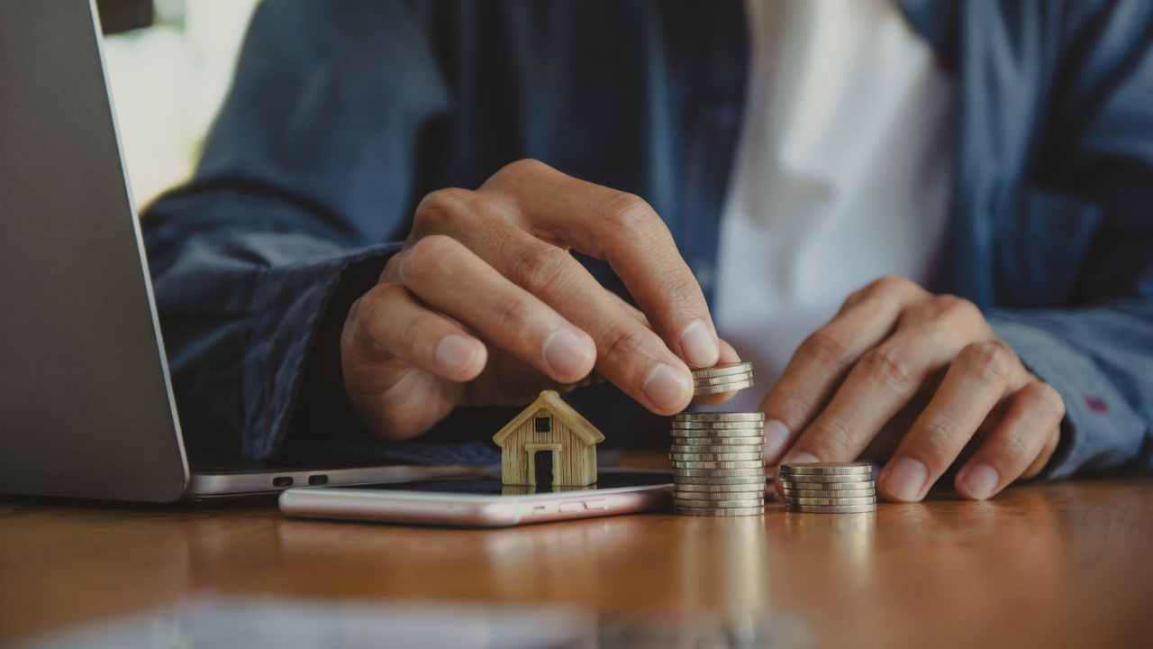 A homeowner stacks coins beside a miniature house on a desk.