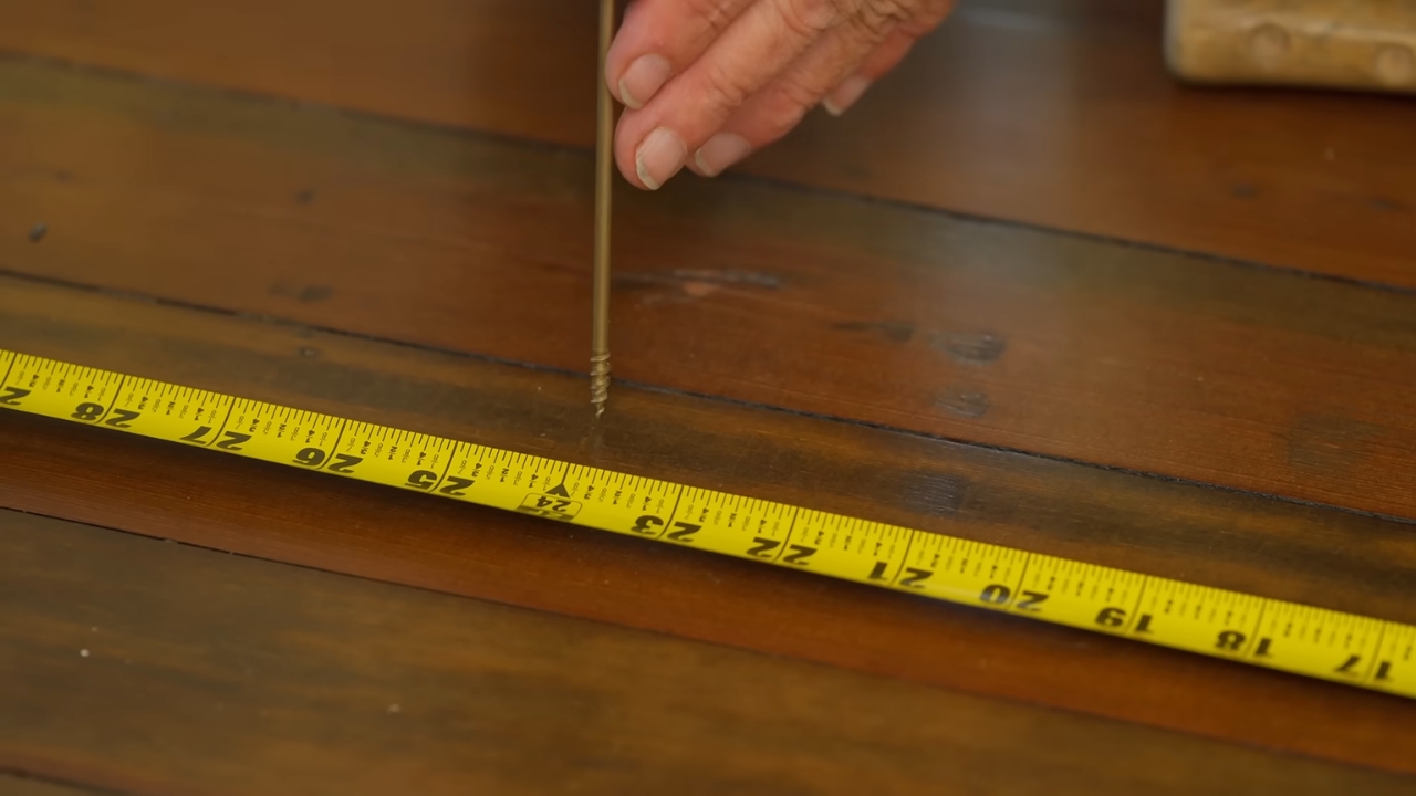 A person positions a screw beside a tape measure on a hardwood floor before a repair.