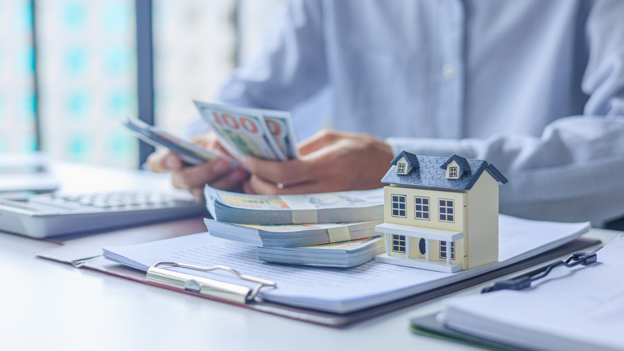 A person counts hundred-dollar bills beside a model house and financial documents.