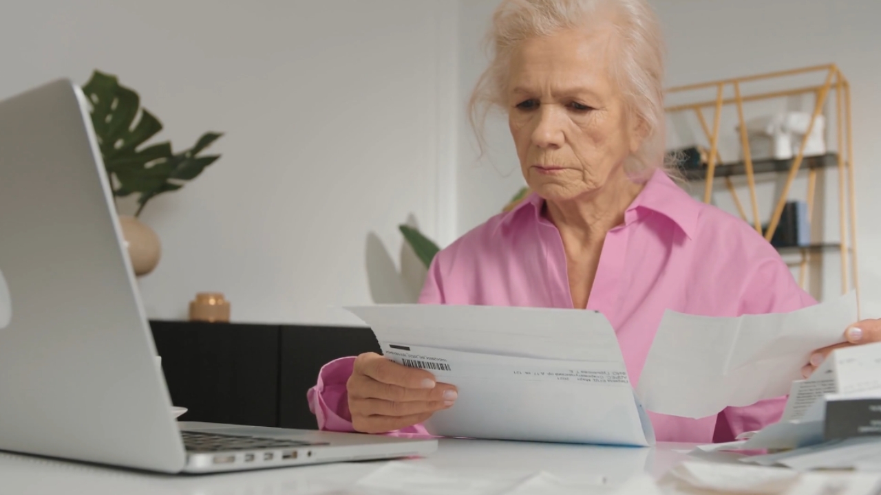An older woman reviews household bills beside her laptop.