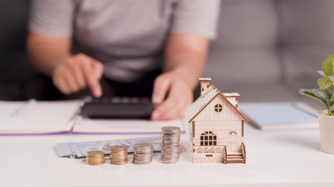 Homeowner calculates costs beside a model house and stacks of coins.