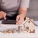 Homeowner calculates costs beside a model house and stacks of coins.