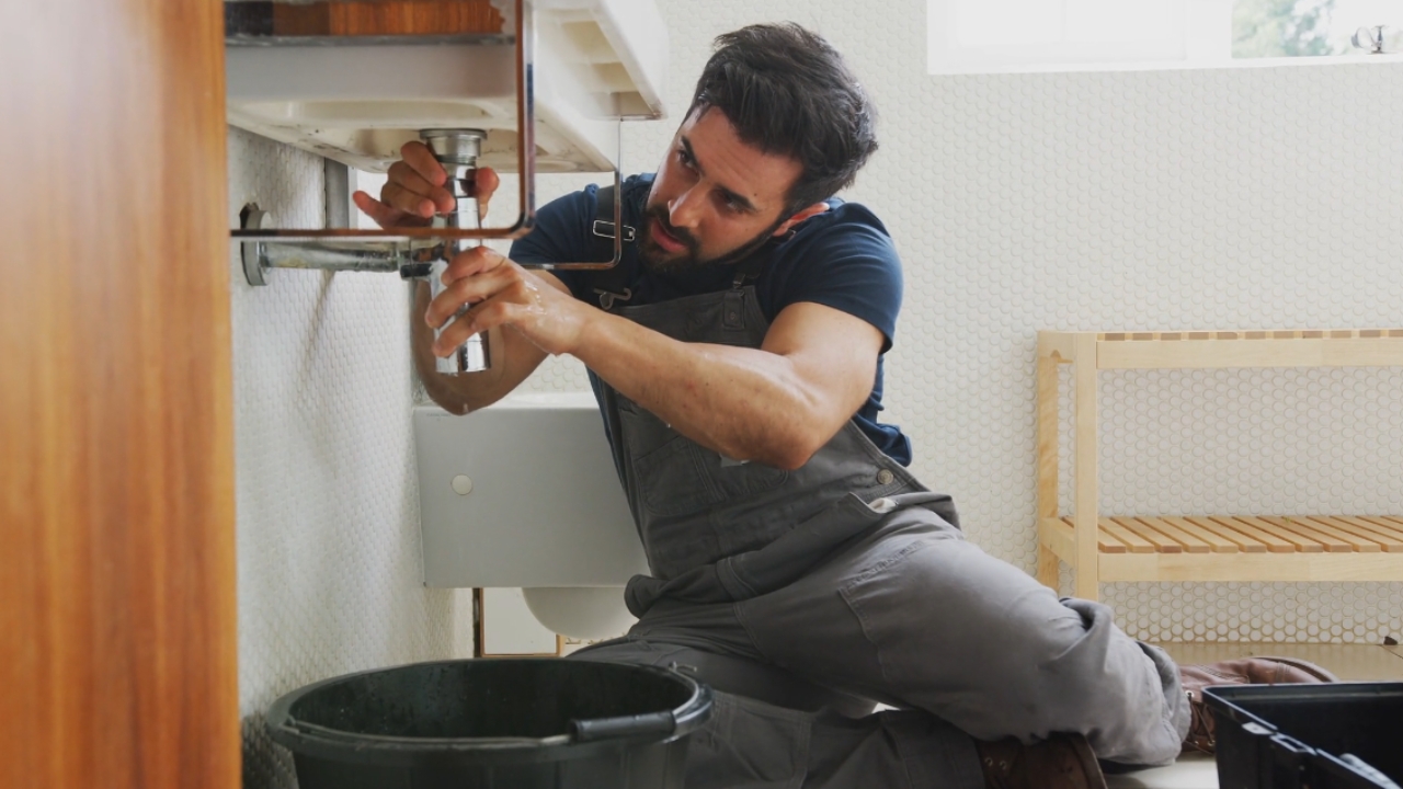 Plumber repairs a pipe beneath a bathroom sink.