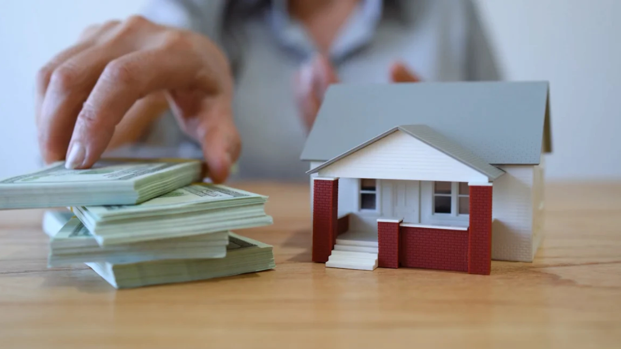 A person reaches for stacks of cash beside a model house on a wooden table.