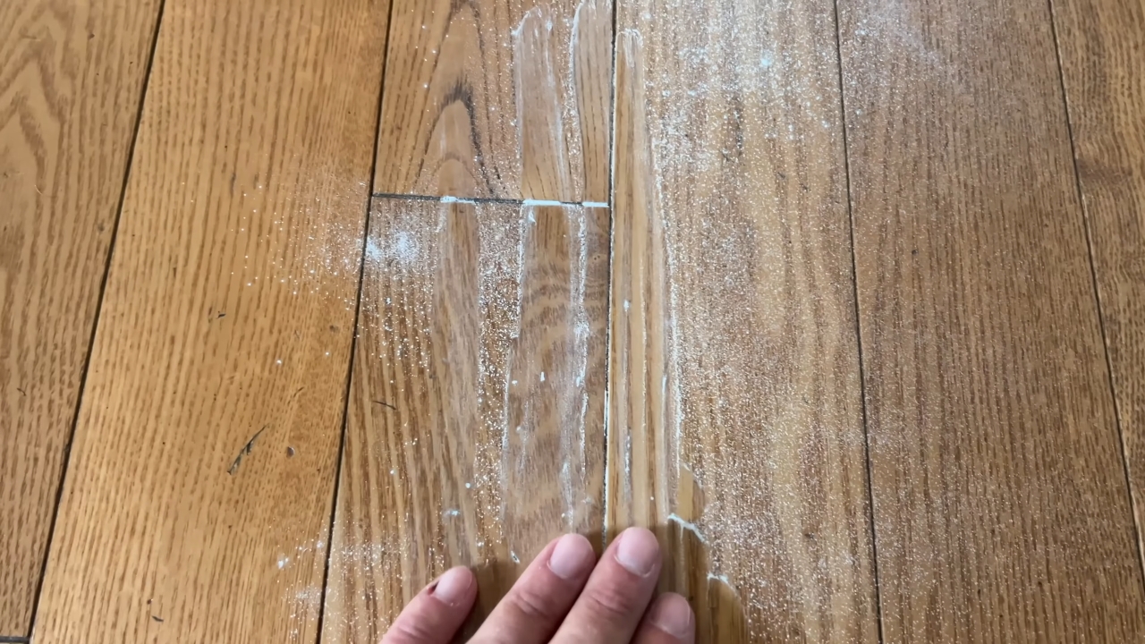 A hand checks a gap between hardwood floorboards covered with white powder.